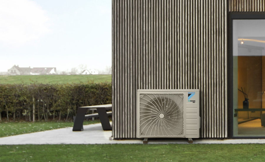 Daikin Sensira buitenunit tegen een houten gevel, met een tuin ernaast